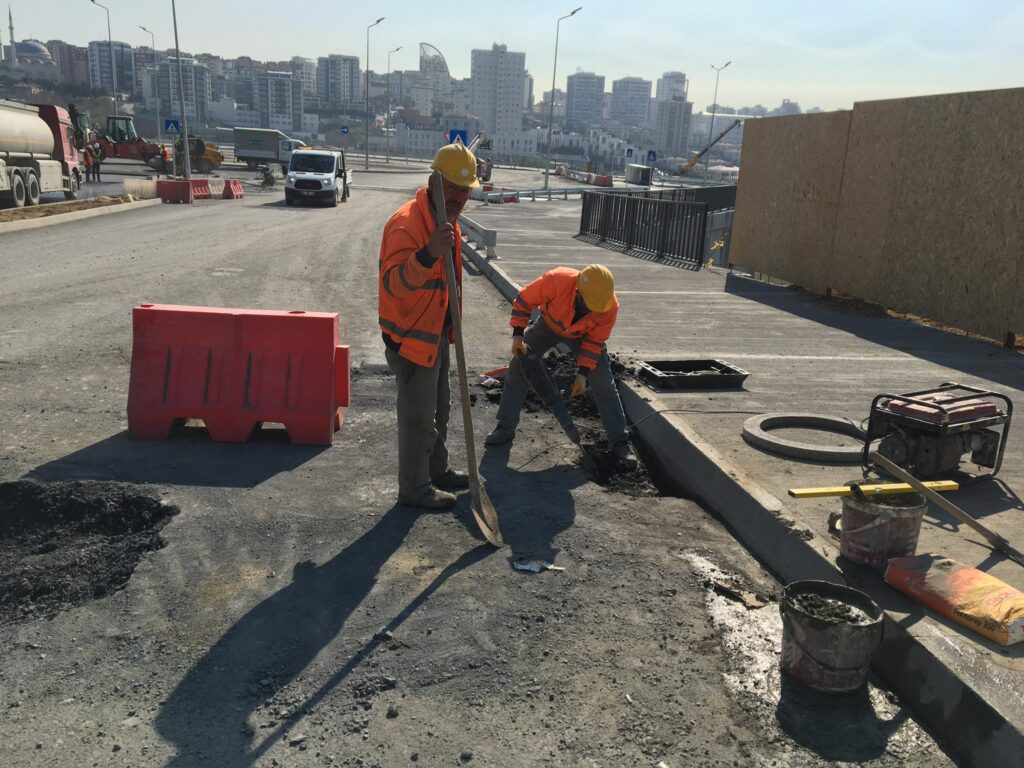 Altyapı çalışması kapsamında yol kenarında asfalt kesim alanı ve beton muayene bacası çevresinde yağmur suyu drenaj hattı montajı yapan işçiler.