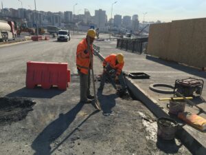 Altyapı çalışması kapsamında yol kenarında asfalt kesim alanı ve beton muayene bacası çevresinde yağmur suyu drenaj hattı montajı yapan işçiler.